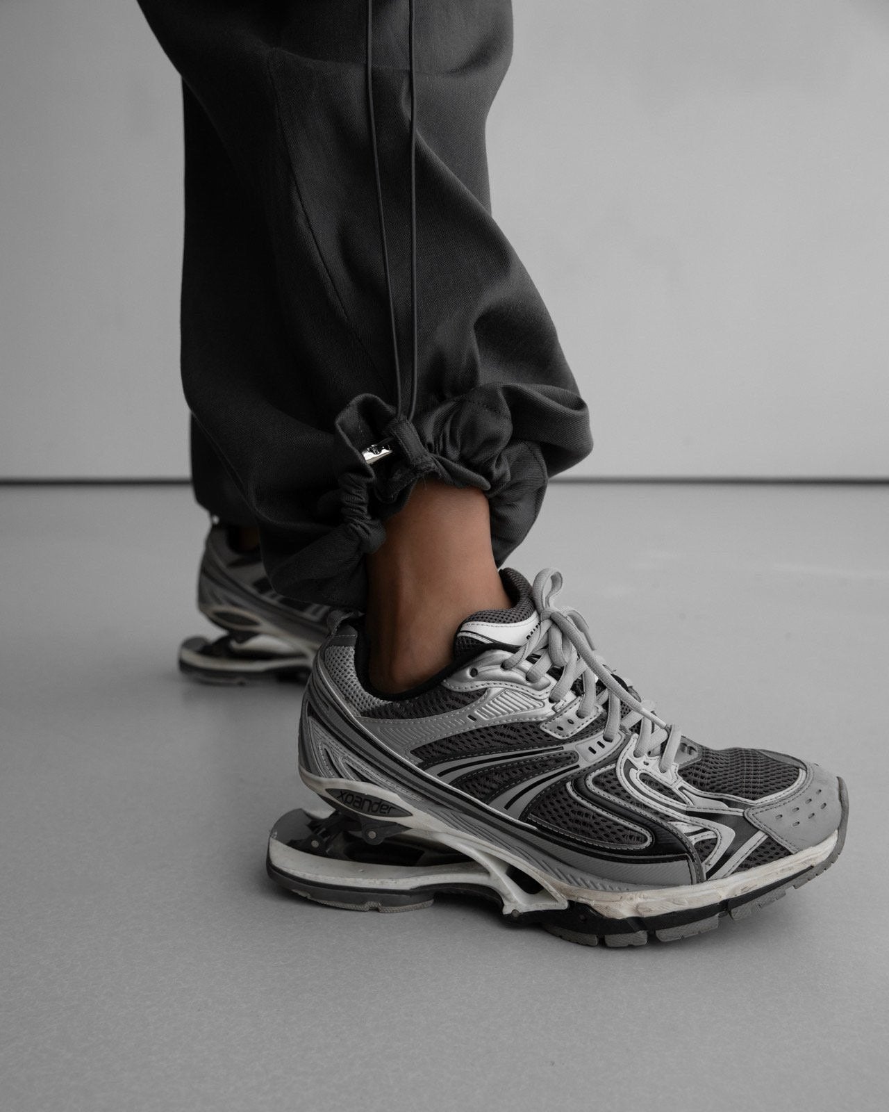 Detail shot of elastic drawstring hem and hardware toggle on the pants, worn with sculptural grey athletic sneakers.
