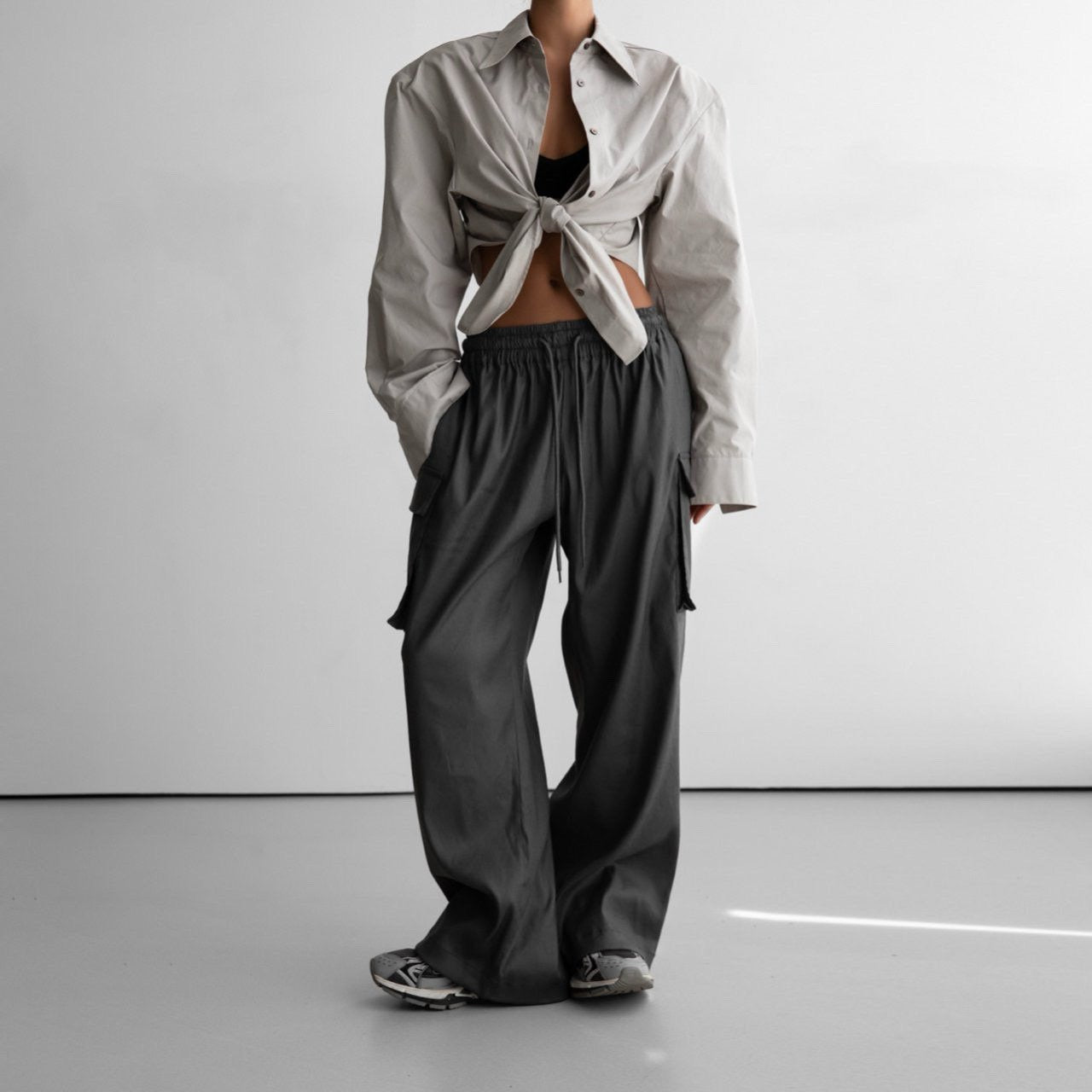 Full body shot of model in dark grey wide-leg cargo pants and tied neutral shirt, styled with casual sneakers in a minimalist setting.