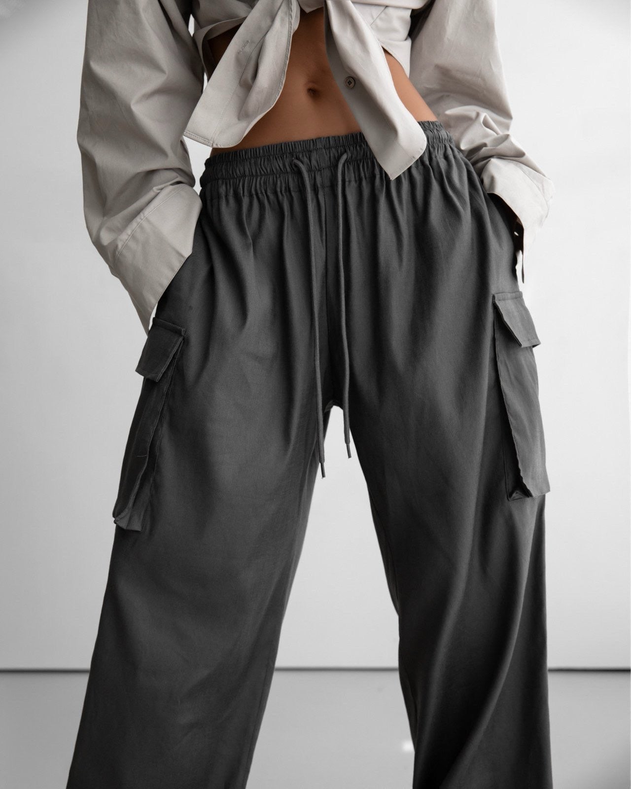 Close-up front view of dark grey wide-leg cargo pants with elastic waistband and side flap pockets, styled with a tied beige shirt.