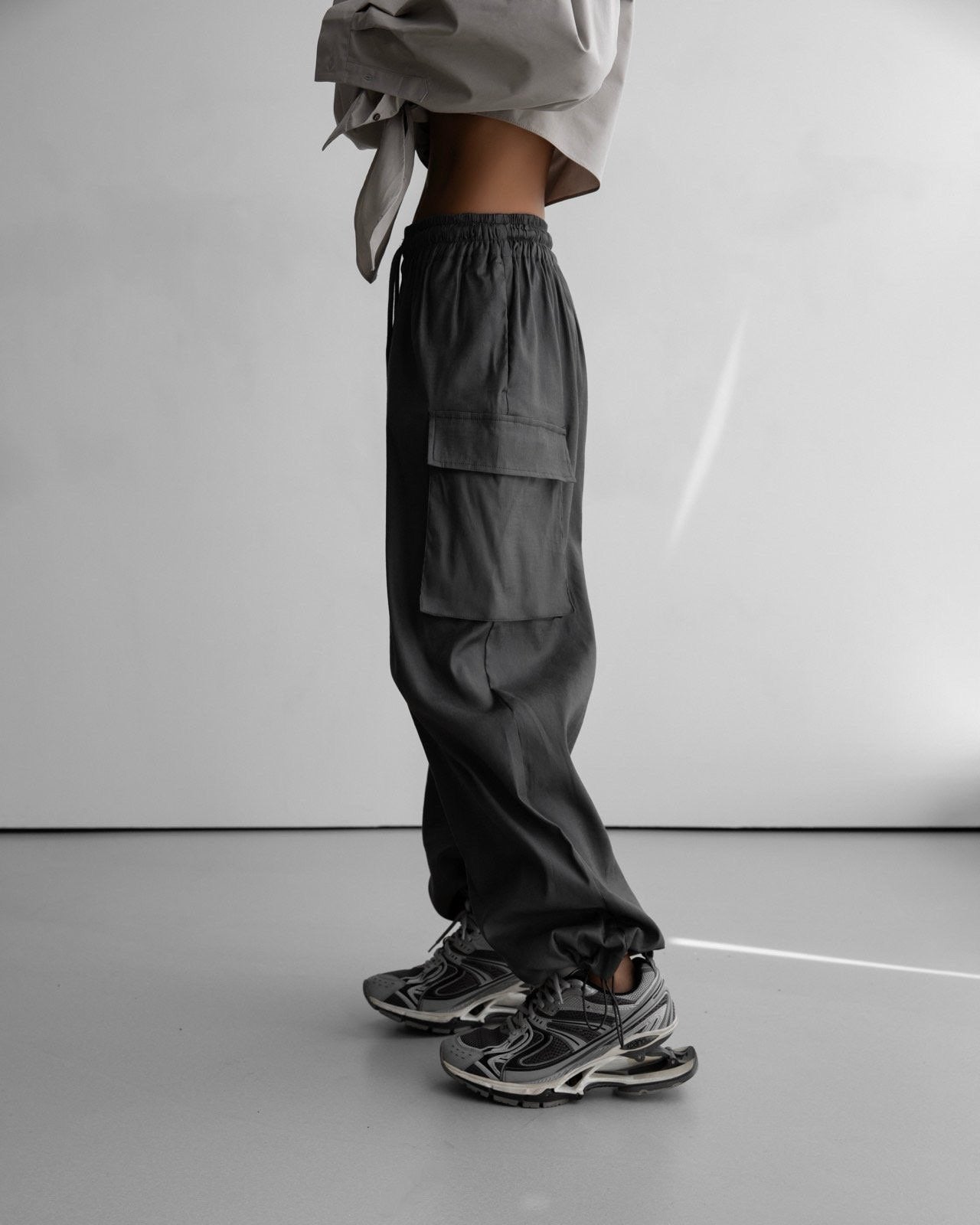 Side profile of dark grey cargo pants with oversized pocket detailing and a relaxed fit, paired with chunky grey sneakers