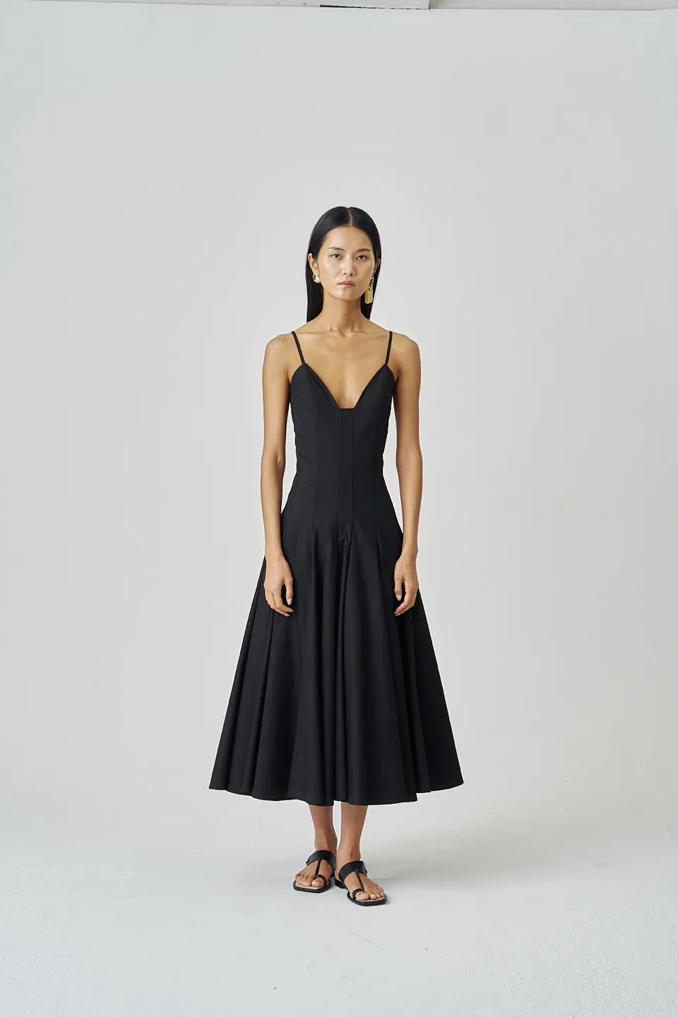 Full-length front view of a woman wearing a flared black midi dress with side pockets and minimal sandals.