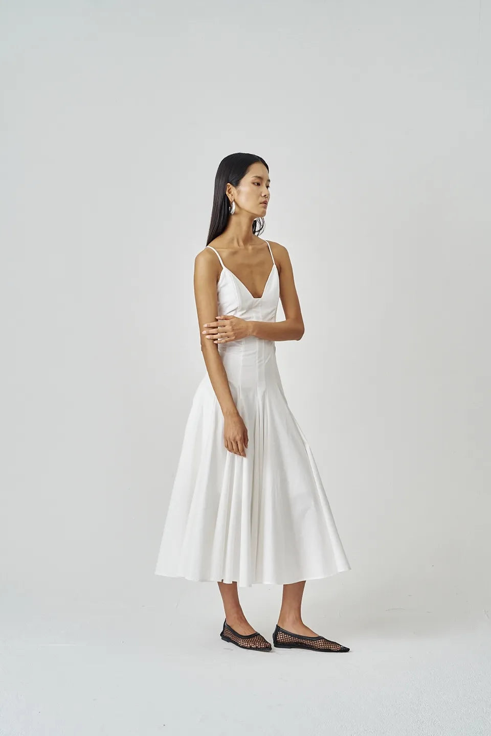 Model wearing white flared midi dress with spaghetti straps, facing left against light background.