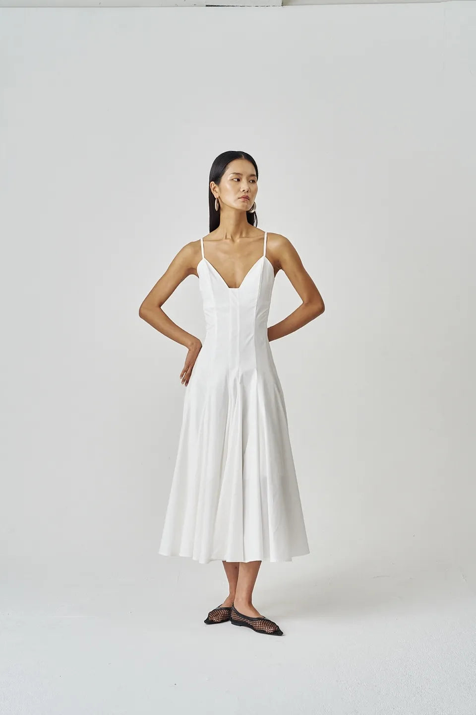 Model standing in white A-line midi dress with thin straps and flared skirt, hands on hips, minimal backdrop.