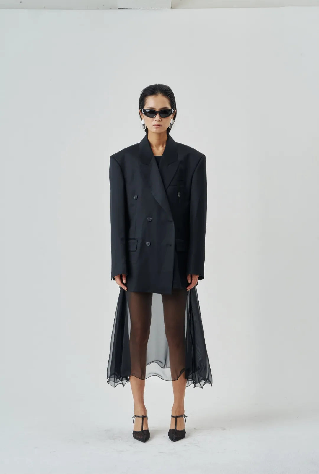 Model wearing a sheer black wire-hem midi skirt layered under an oversized black blazer with structured shoulders and heels