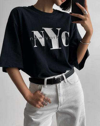 Oversized navy NYC graphic t-shirt styled with white pants and black belt, shot from waist up