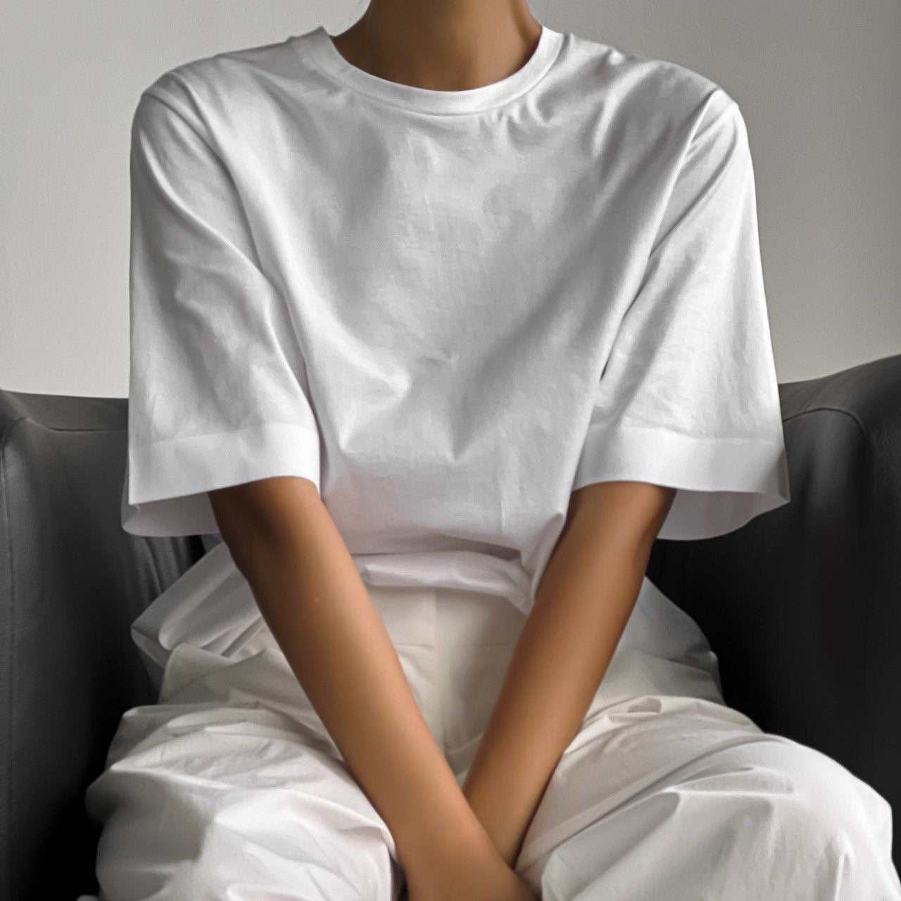 Front-facing view of a woman in a structured white t-shirt and relaxed tailored white pants, seated with arms gently crossed.
