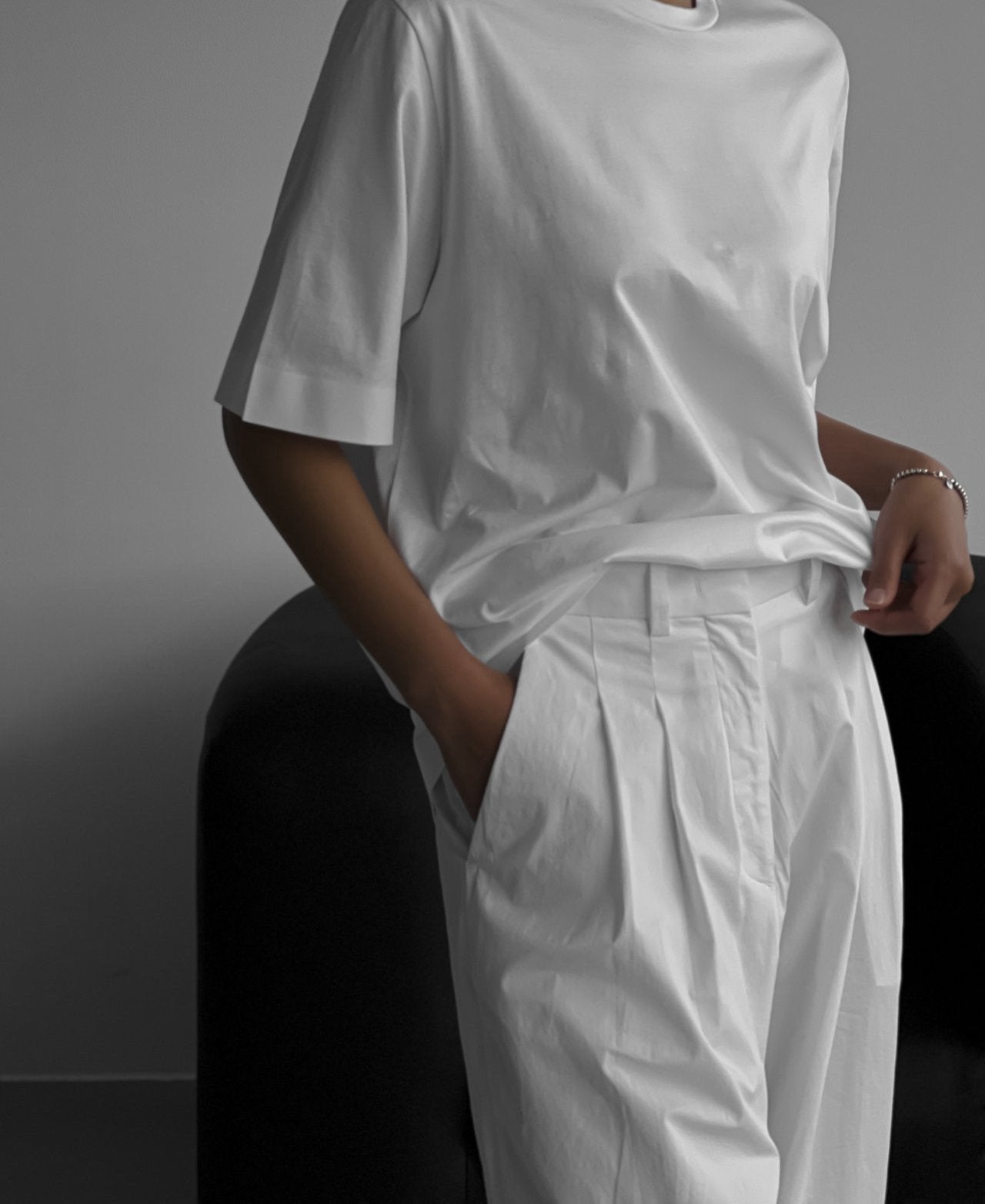 Close-up of a woman wearing an oversized crisp white t-shirt tucked into pleated white trousers, seated on a dark armchair.