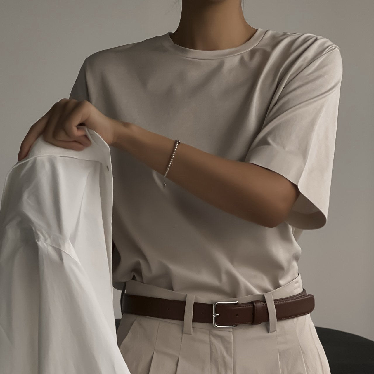 Minimalist light beige crewneck t-shirt styled with pleated trousers and dark brown belt, photographed in soft indoor lighting.