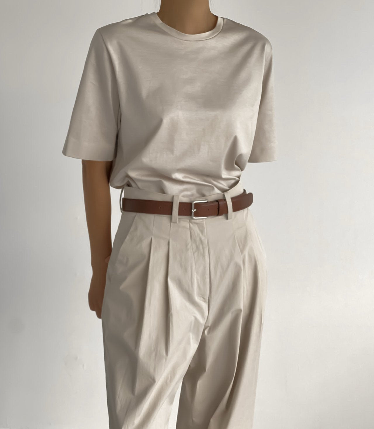 Front view of a sleek, oversized light beige t-shirt tucked into tailored pleated pants, styled with a chocolate brown belt.