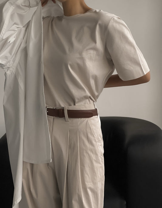 Muted beige t-shirt styled with pleated pants and brown belt, layered with a crisp white shirt in hand, captured in low contrast lighting.