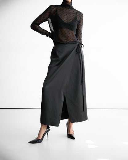 Model in black wrap skirt with sheer top, hands together, minimalist studio backdrop