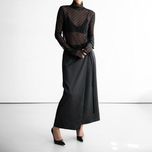 Model wearing black wrap skirt and sheer polka dot top, standing confidently in heels