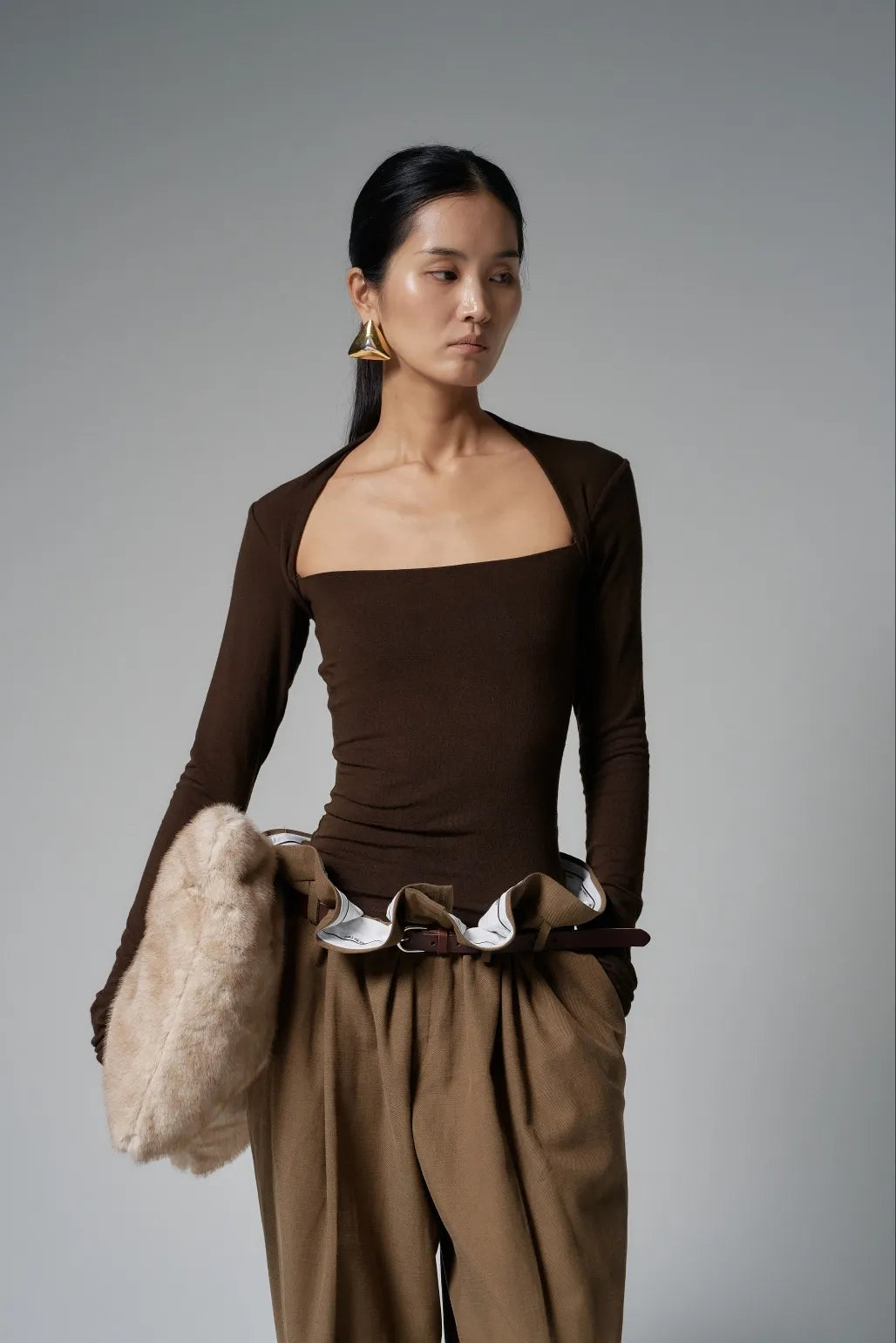 Model wearing the top in chocolate brown, paired with wide tailored trousers, side view holding a faux-fur bag