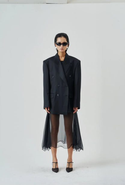 Model wearing a sheer black wire-hem midi skirt layered under an oversized black blazer with structured shoulders and heels