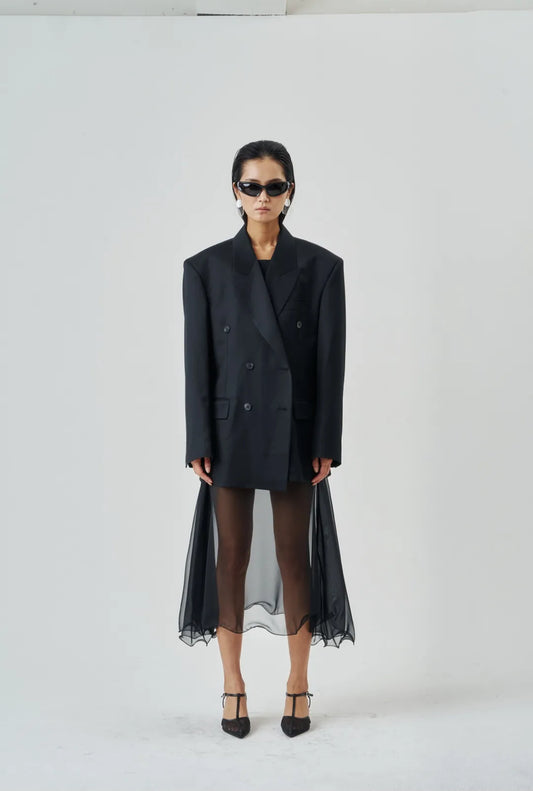 Model wearing a sheer black wire-hem midi skirt layered under an oversized black blazer with structured shoulders and heels