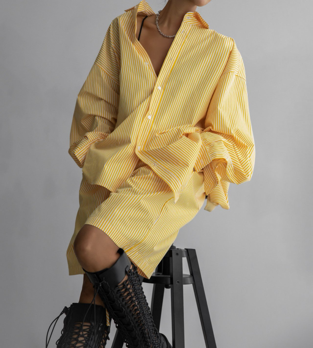 Seated model in yellow striped shorts and oversized shirt, styled with chain necklace and lace-up boots.
